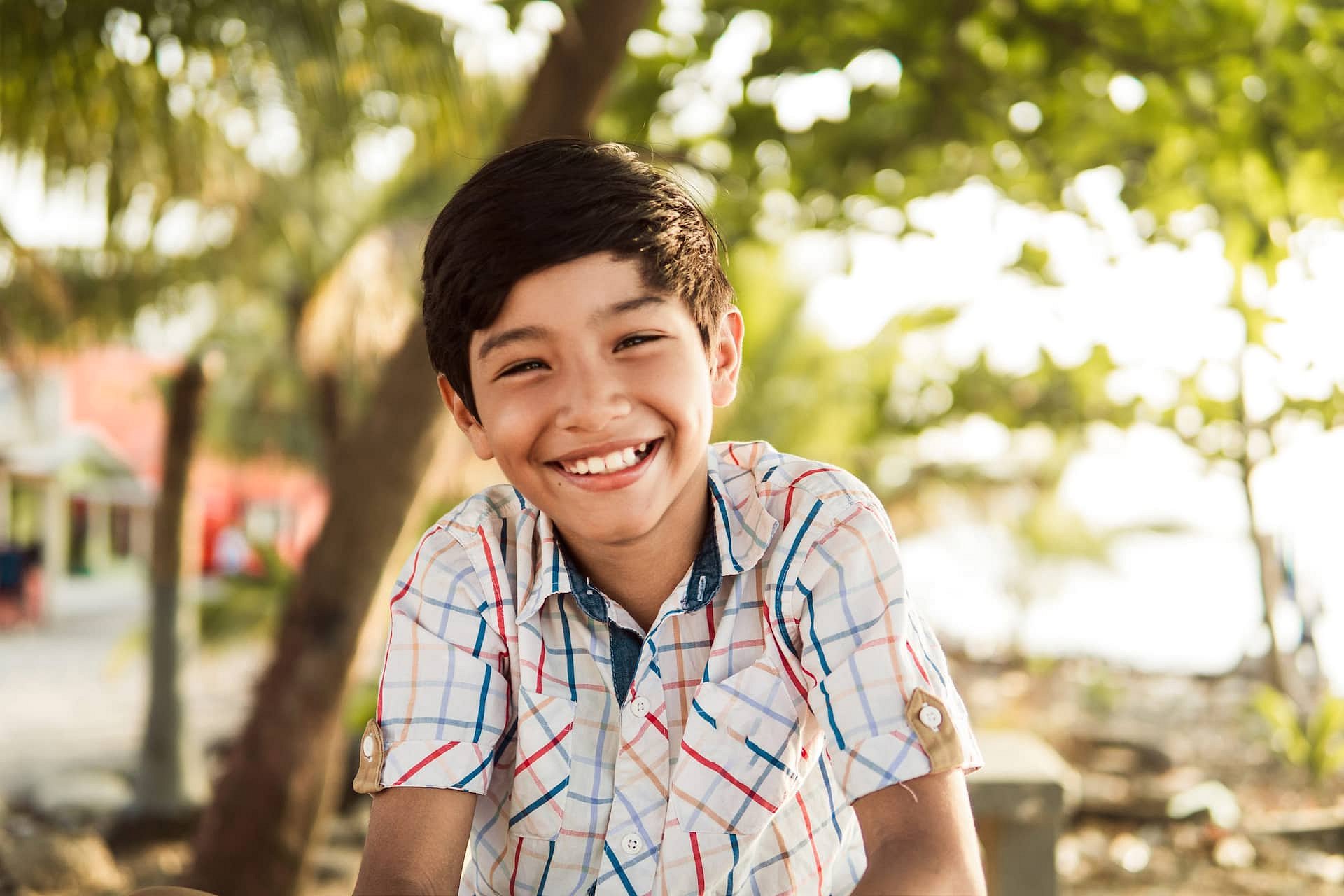 A boy with dark hair in a plaid shirt smiles outdoors amid trees and sunlight, reflecting the confidence that Smile City Orthodontics in Sunnyvale, TX can inspire as your trusted Children’s Orthodontist.