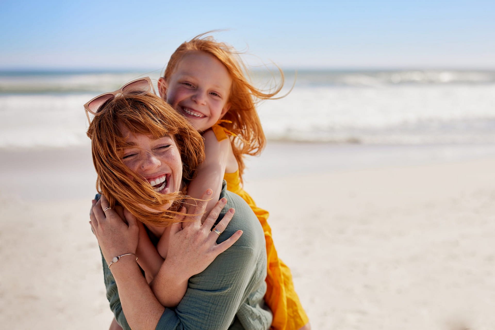 Celebrating a successful jaw surgery, an adult and child with red hair share a joyful piggyback ride on a sunny beach with waves in the background—an experience made possible by Smile City Orthodontics in Sunnyvale, TX.