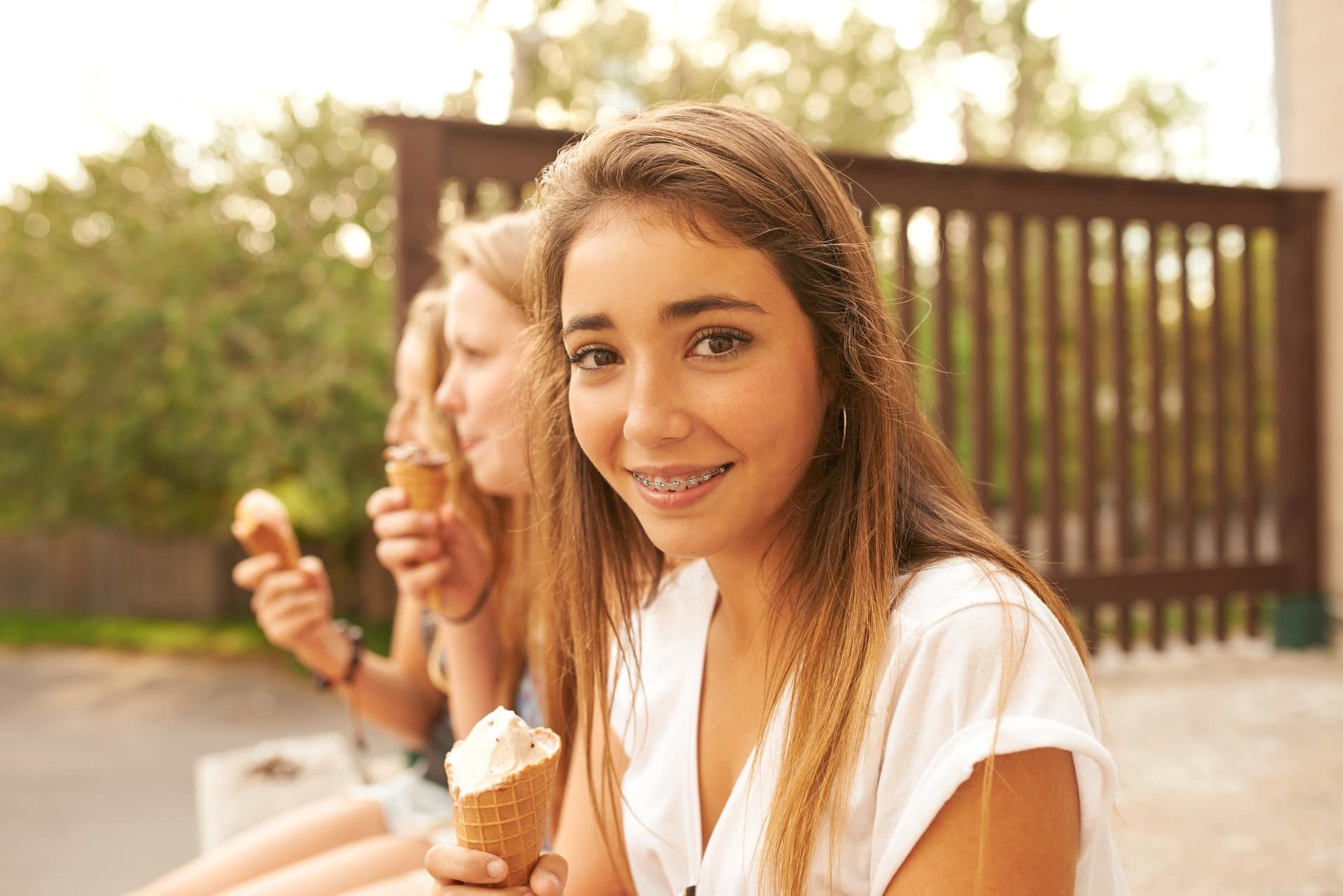 Three teenagers enjoy ice cream cones outdoors; the girl in the foreground, sporting braces and long brown hair, smiles at the camera with a hint of tongue thrust a moment perfectly captured by Smile City Orthodontics in Sunnyvale, TX.
