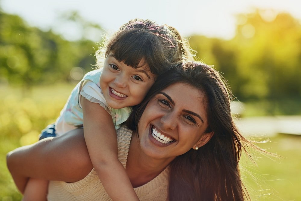 Showcasing bright smiles that could be achieved with Affordable Invisalign, a woman gives a young girl a piggyback ride in a sunlit park—capturing the happy results possible at Smile City Orthodontics in Sunnyvale, TX.