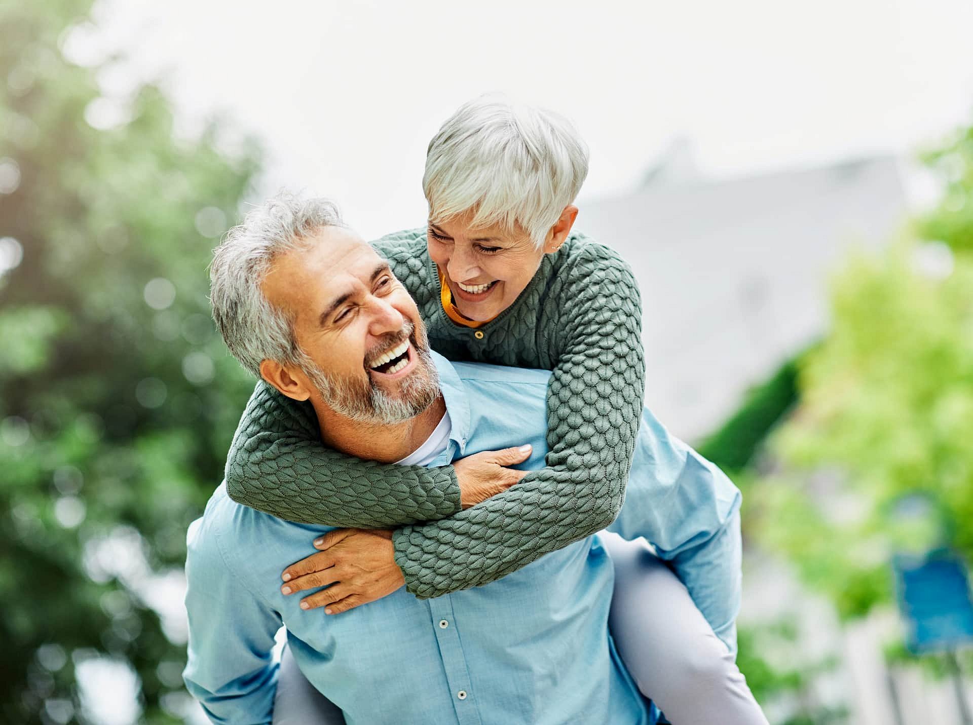 A joyful older couple shares a piggyback ride outdoors, both smiling confidently after jaw surgery at Smile City Orthodontics in Sunnyvale TX, with trees and greenery in the background.