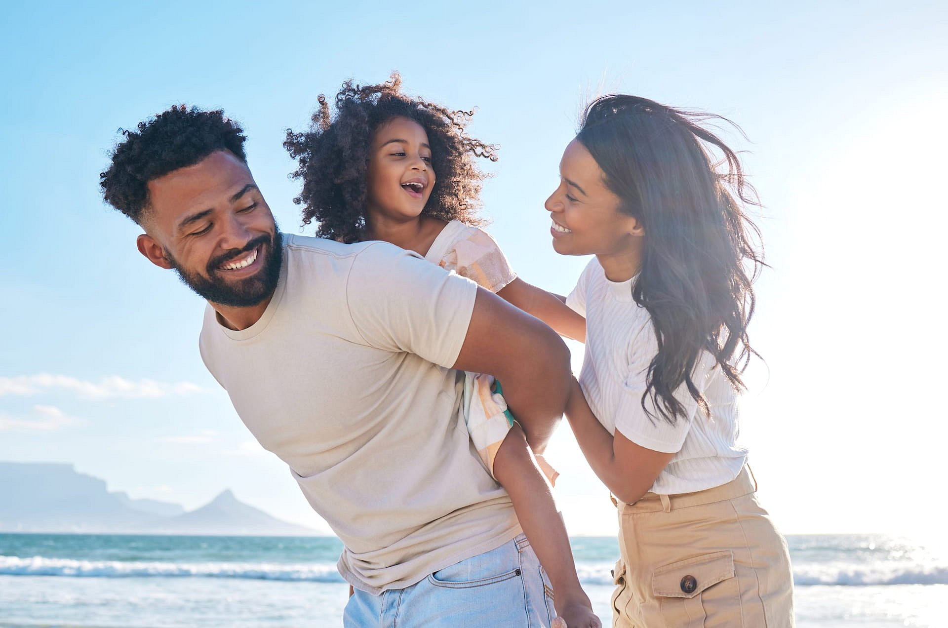 At Smile City Orthodontics in Sunnyvale TX, a man gives a piggyback ride to a smiling child while a woman stands beside them on a sunny beach—reminding families to cherish every moment and smile confidently with our Free Orthodontic Consultation.