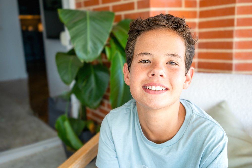 At Smile City Orthodontics in Sunnyvale TX, a young boy in a light blue shirt smiles near a green plant and brick wall, possibly considering whether Invisalign is covered by insurance for his future smile.