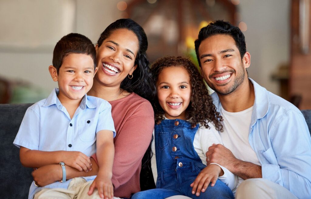 At Smile City Orthodontics in Sunnyvale TX, a family of four—two adults and two children—sits together on a well-lit couch, smiling at the camera.