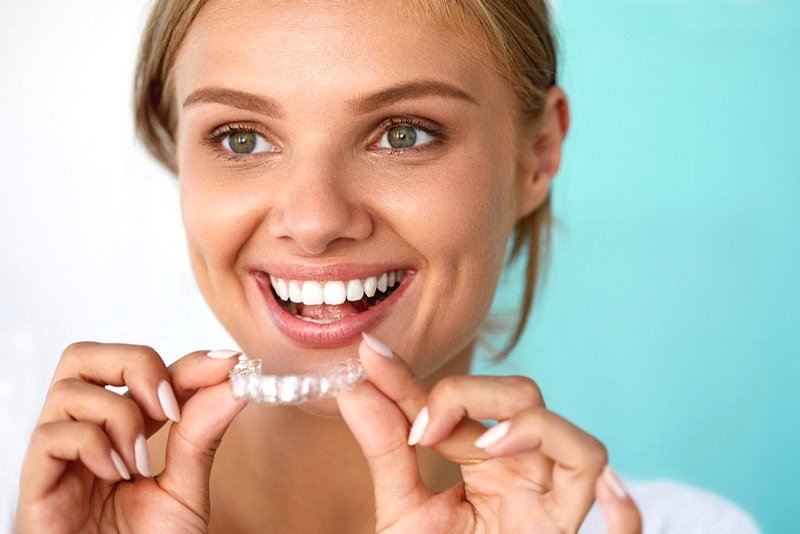 A smiling woman in front of a light blue background holds a clear dental aligner, showcasing confidence and the benefits of orthodontic insurance plans at Smile City Orthodontics in Sunnyvale, TX.