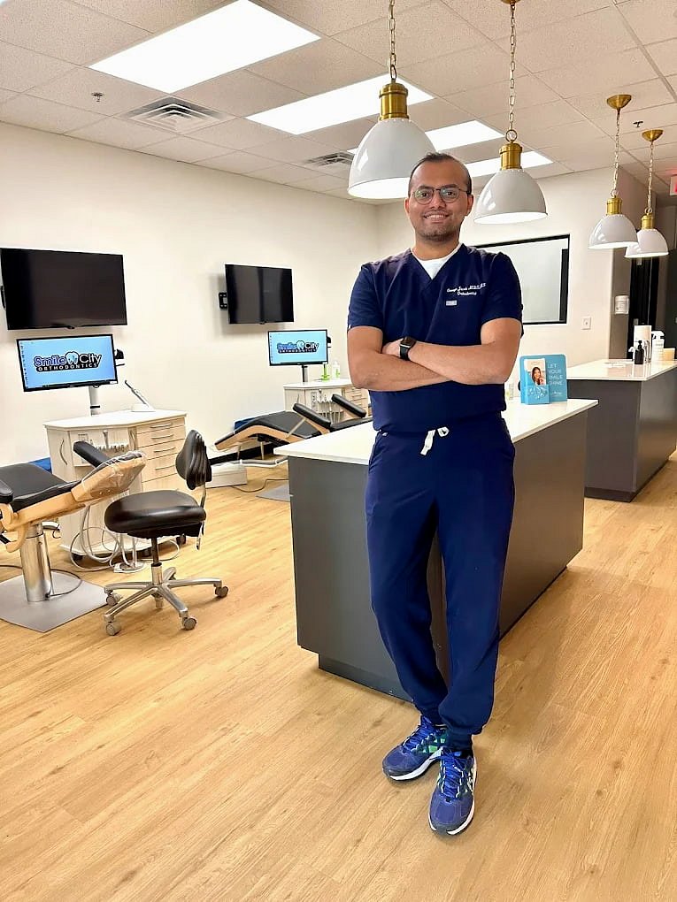 At Smile City Orthodontics in Mesquite TX, Dr. Jacob, BDS, MDS, MS an expert orthodontist, stands confidently with arms crossed in a modern office featuring dental chairs, monitors, and bright lighting.