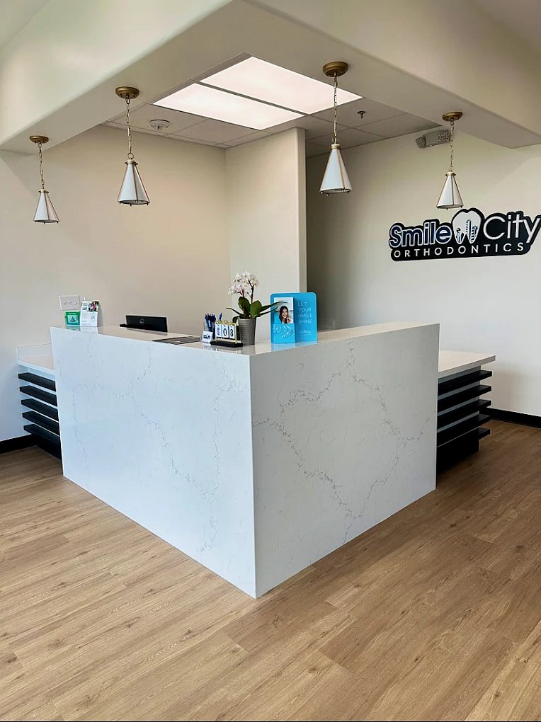 Featuring a modern orthodontic reception desk with a marble finish, pendant lighting, wood flooring, and a Smile City Orthodontics sign on the wall, this space in Sunnyvale, TX offers a welcoming atmosphere.