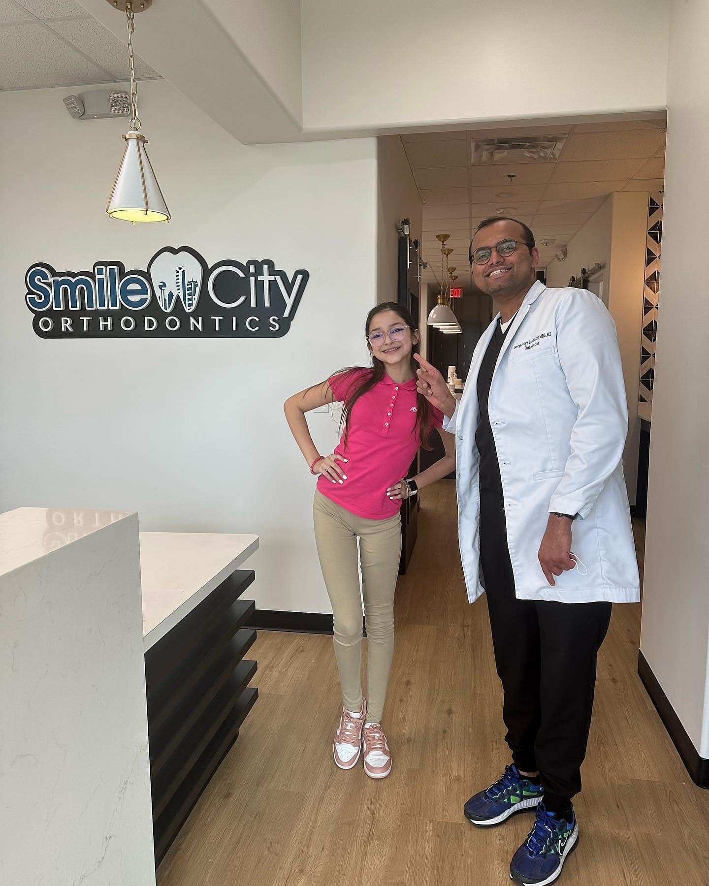 Inside an office in Sunnyvale, TX, a smiling young girl stands beside an orthodontist in a white coat, with a "Smile City Orthodontics" sign visible on the wall.