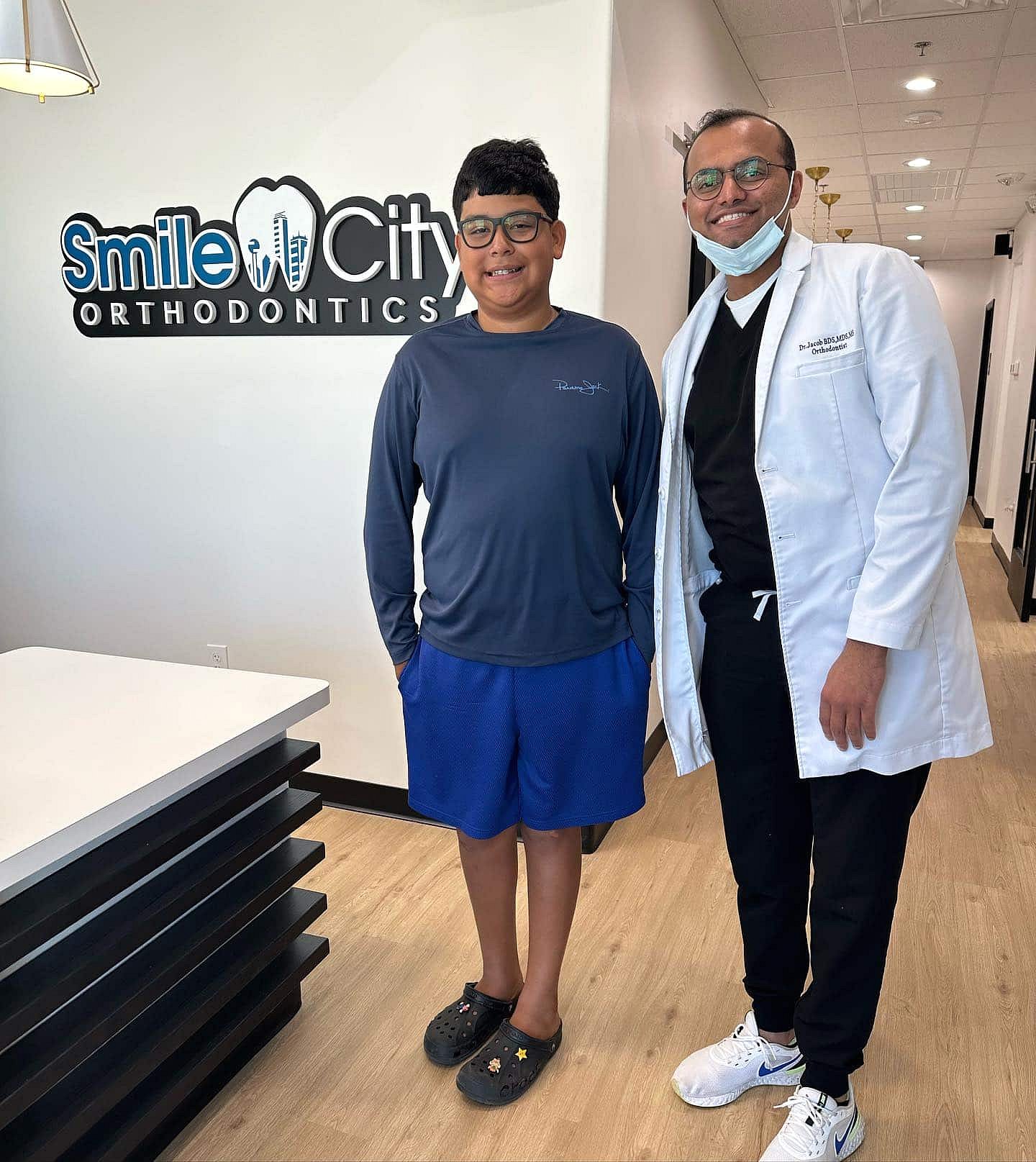 Inside Smile City Orthodontics in Sunnyvale, TX, a boy stands with an orthodontist in front of the clinic's logo displayed on the wall.