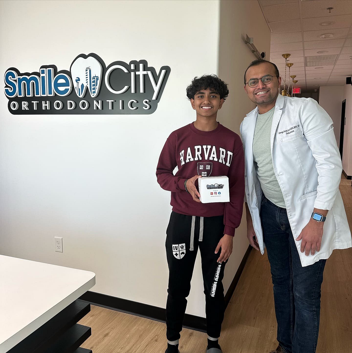 At Smile City Orthodontics in Sunnyvale, TX, a young person in a Harvard sweatshirt stands beside a smiling orthodontist, holding a small white box.