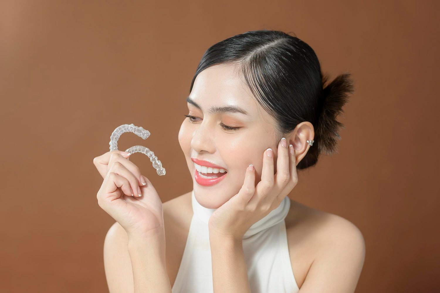 A woman smiles, holding Invisalign® Clear Aligners in one hand and resting her other hand on her face against a plain brown background at Smile City Orthodontics in Mesquite, TX.
