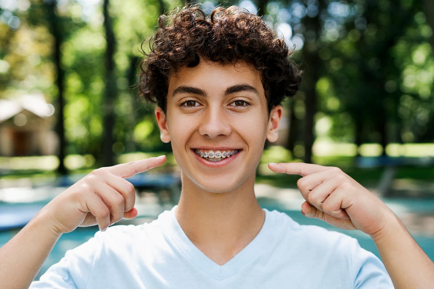 A teenage boy with curly hair smiles and points to his braces with both hands outdoors, with trees and blurred greenery in the background at Smile City Orthodontics in Sunnyvale, TX.
