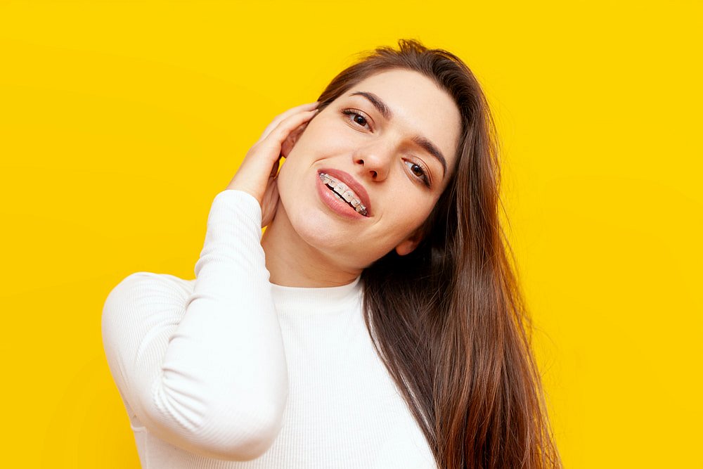 A woman with long brown hair, wearing a white top and clear braces, smiles and touches her neck against a bright yellow background at Smile City Orthodontics in Mesquite, TX.