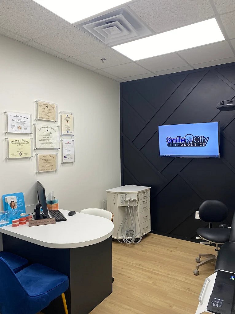 In Sunnyvale, TX, Smile City Orthodontics features a modern office equipped with a desk, computer, dental chair, wall-mounted TV displaying their name, and framed certificates.