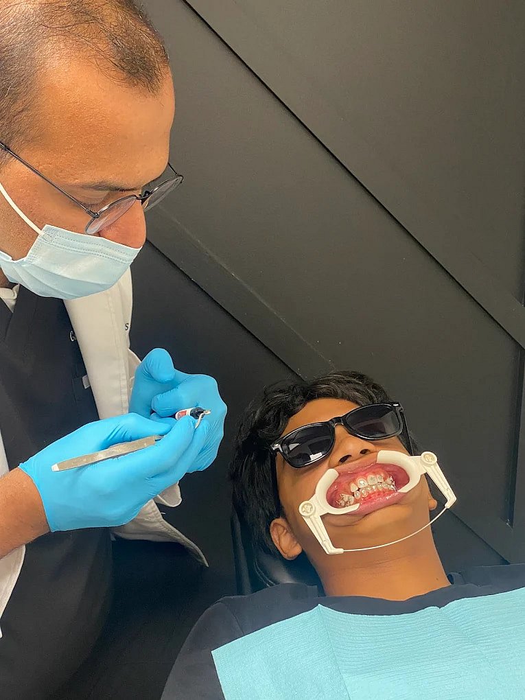 At Smile City Orthodontics in Mesquite TX, a dentist with gloves and a mask examines a patient's braces as the patient wears sunglasses and a cheek retractor.