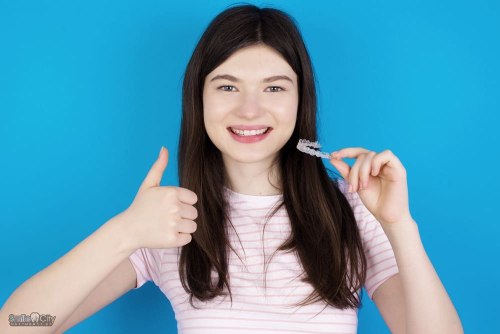 Dark-haired girl giving a thumbs up while holding a clear aligner and smiling after understanding Does Insurance Cover Invisalign in Sunnyvale, TX