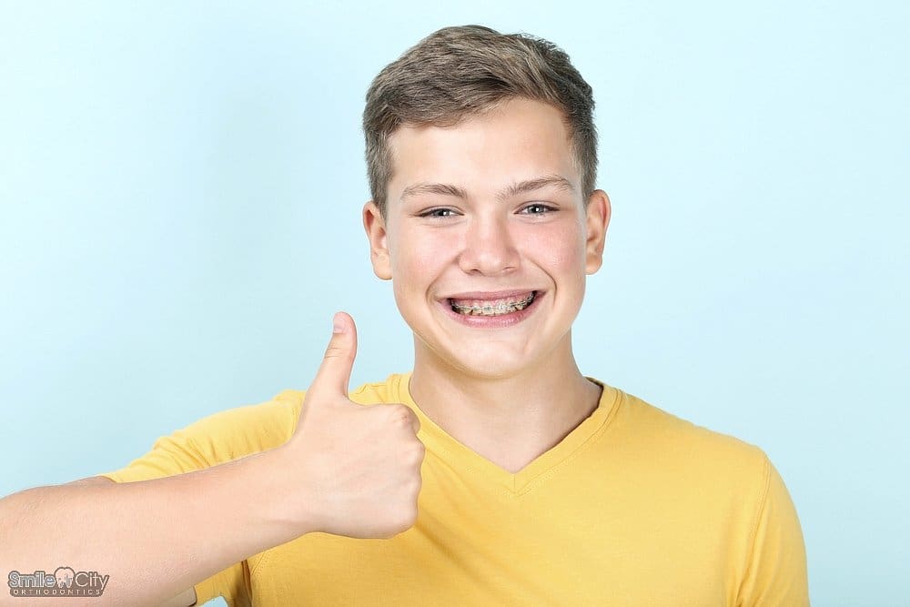young man with metal braces showing thumb up on blue background - How to Get Braces Glue Off Teeth in Sunnyvale, TX