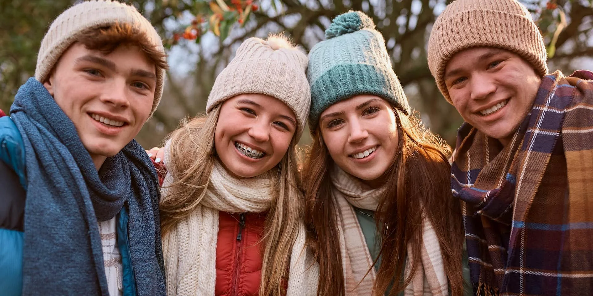 A group of four friends wearing winter hats, scarves, and jackets stands close together outdoors, smiling at the camera with autumn trees behind them, discussing everything from weekend plans to Invisalign cost at Smile City Orthodontics in Sunnyvale, TX.