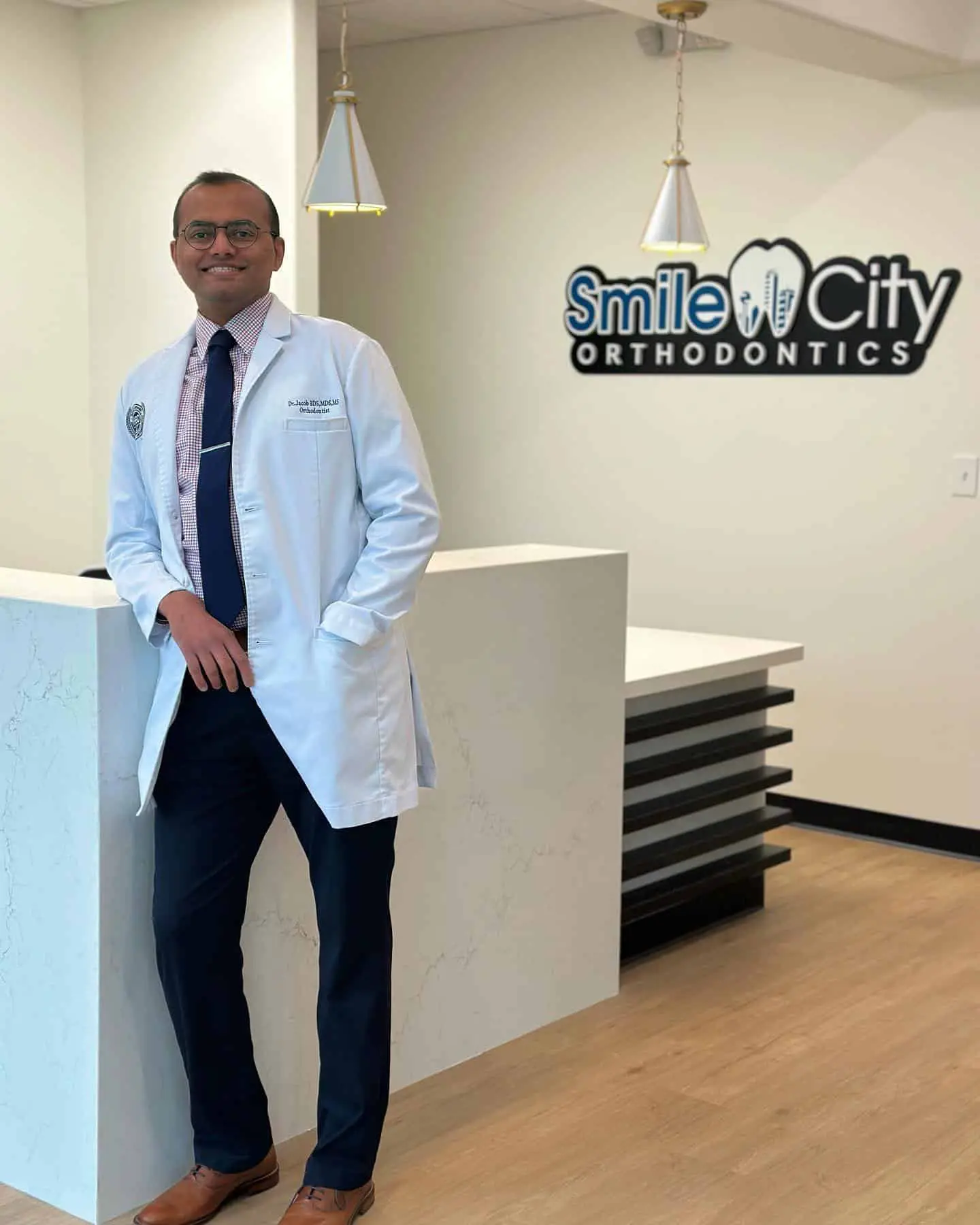 At Smile City Orthodontics in Sunnyvale TX, Dr. Jacob, an orthodontics expert in a white lab coat, stands by the reception desk with the clinic’s sign visible behind him.